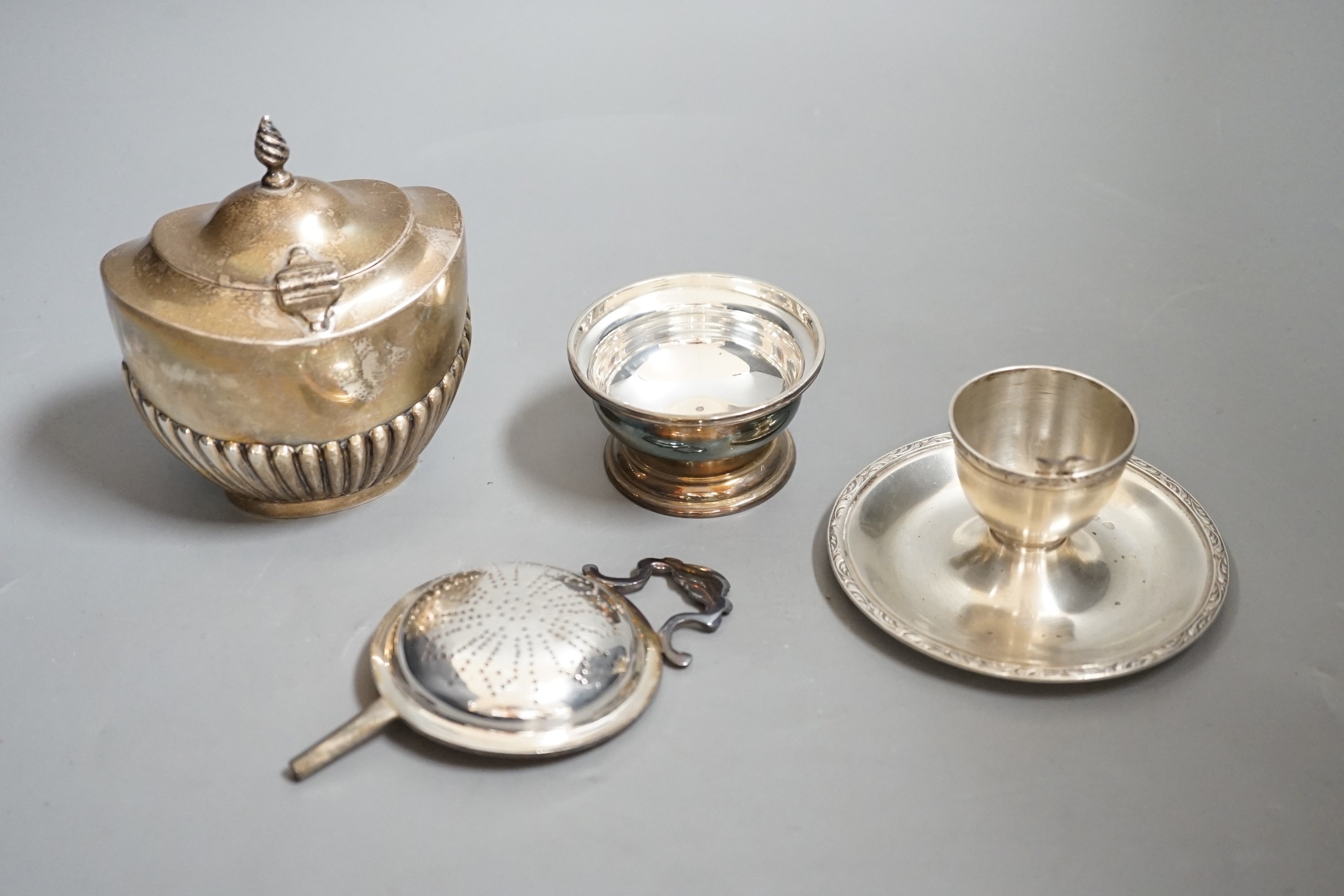 A late Victorian silver half-fluted tea caddy, Birmingham, 1900, a silver strainer on stand, a silver egg cup stand and a pair of silver server spoons.